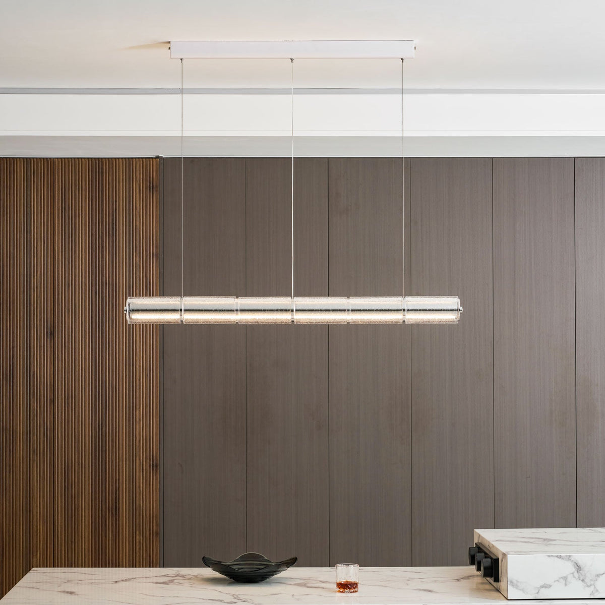 A Clérachic linear chandelier of clear glass segments glowing above a marble dining counter, with a dark bowl and a tumbler below.