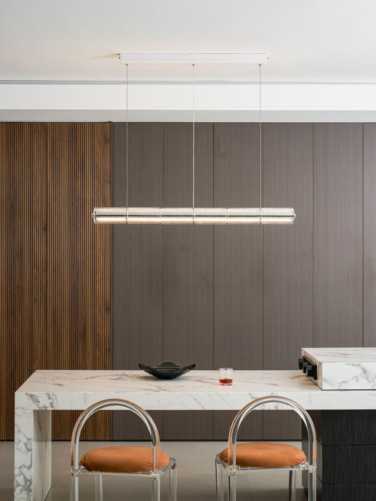 Compact Clérachic glass chandelier lit above a marble breakfast bar with two clear-backed stools and orange cushions.