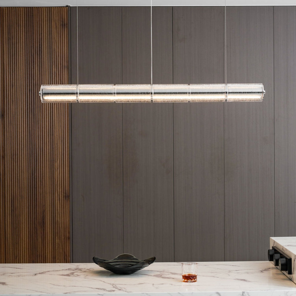 Close-up room view of a long Clérachic glass chandelier with warm light, against a dark wood wall above a marble counter.