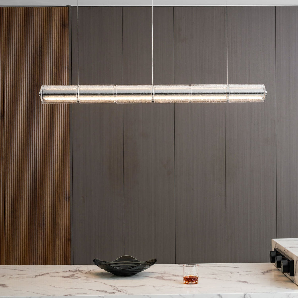 Close-up room view of a long Clérachic glass chandelier with warm light, against a dark wood wall above a marble counter.