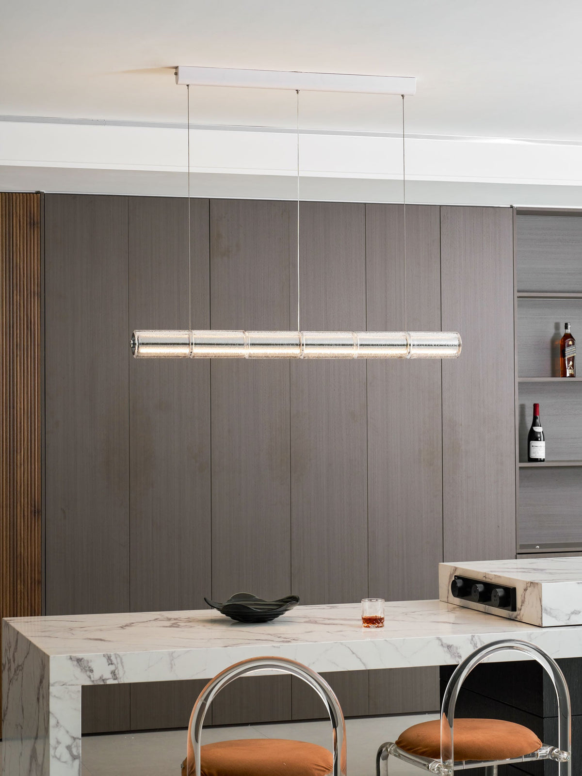 Clérachic segmented glass chandelier lit above a marble island, with wine bottles on shelves in the dark panelled wall behind.