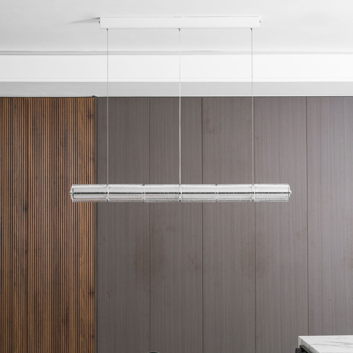 Unlit chrome and clear glass chandelier from Clérachic hung against a grey-brown panelled wall, with a ribbed walnut panel on the left.