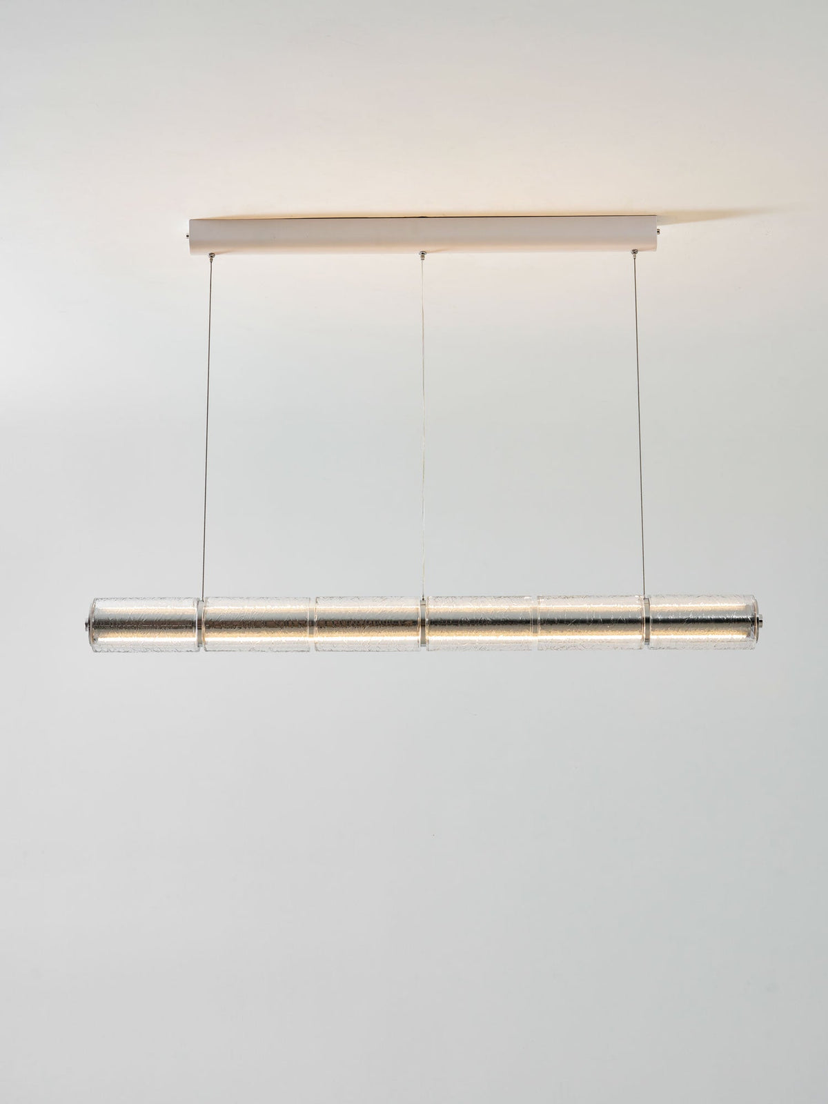 Lit Clérachic glass chandelier hanging from a white ceiling canopy on three cables, with warm light grazing the ceiling above.