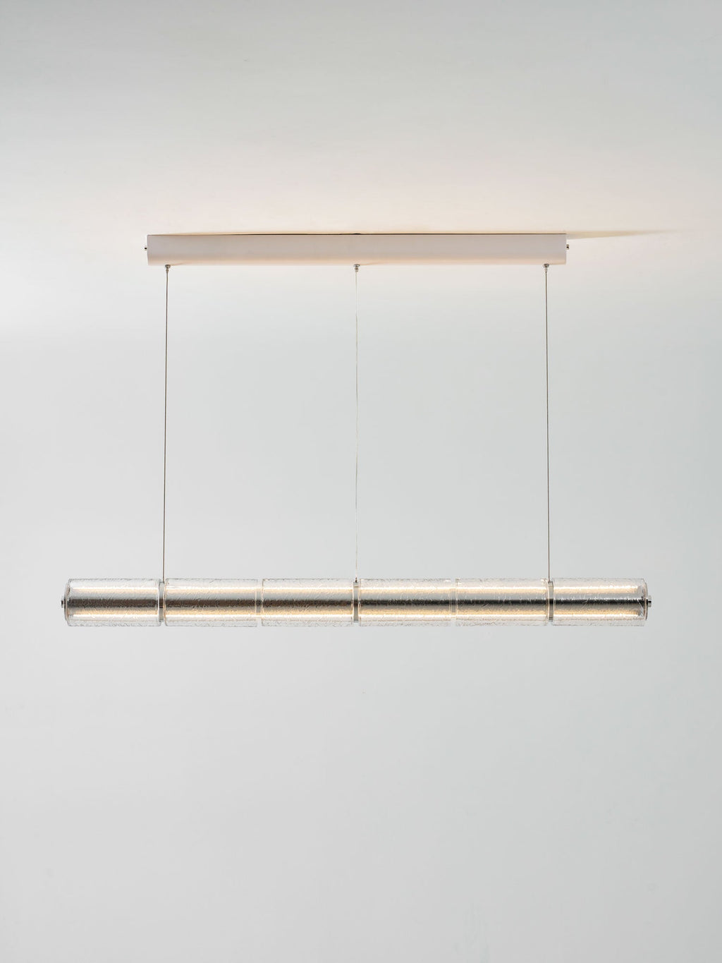 Lit Clérachic segmented glass chandelier with a soft warm glow, hung from a slim ceiling canopy against a light grey wall.