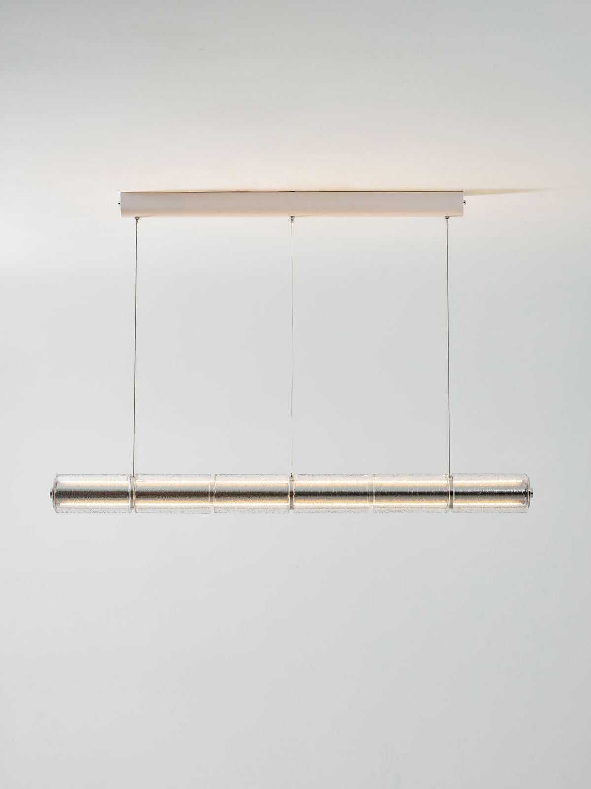 Lit Clérachic segmented glass chandelier with a soft warm glow, hung from a slim ceiling canopy against a light grey wall.
