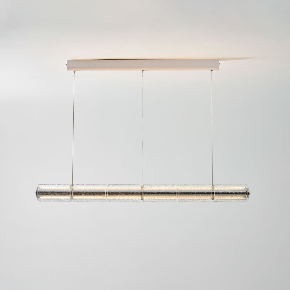Warm-lit Clérachic glass chandelier hanging from a chrome ceiling bar, light spreading across the pale wall behind it.