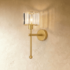 Clérachic all-gold wall lamp with a clear ribbed glass drum shade, a slim stem and ball finial, glowing on a limewash wall.