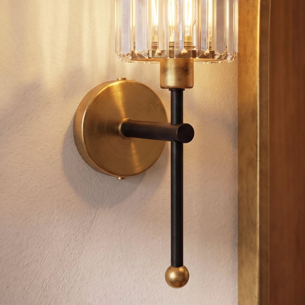 Clérachic close view of a black stem, brushed brass backplate and ball finial under a clear ribbed glass shade on plaster.