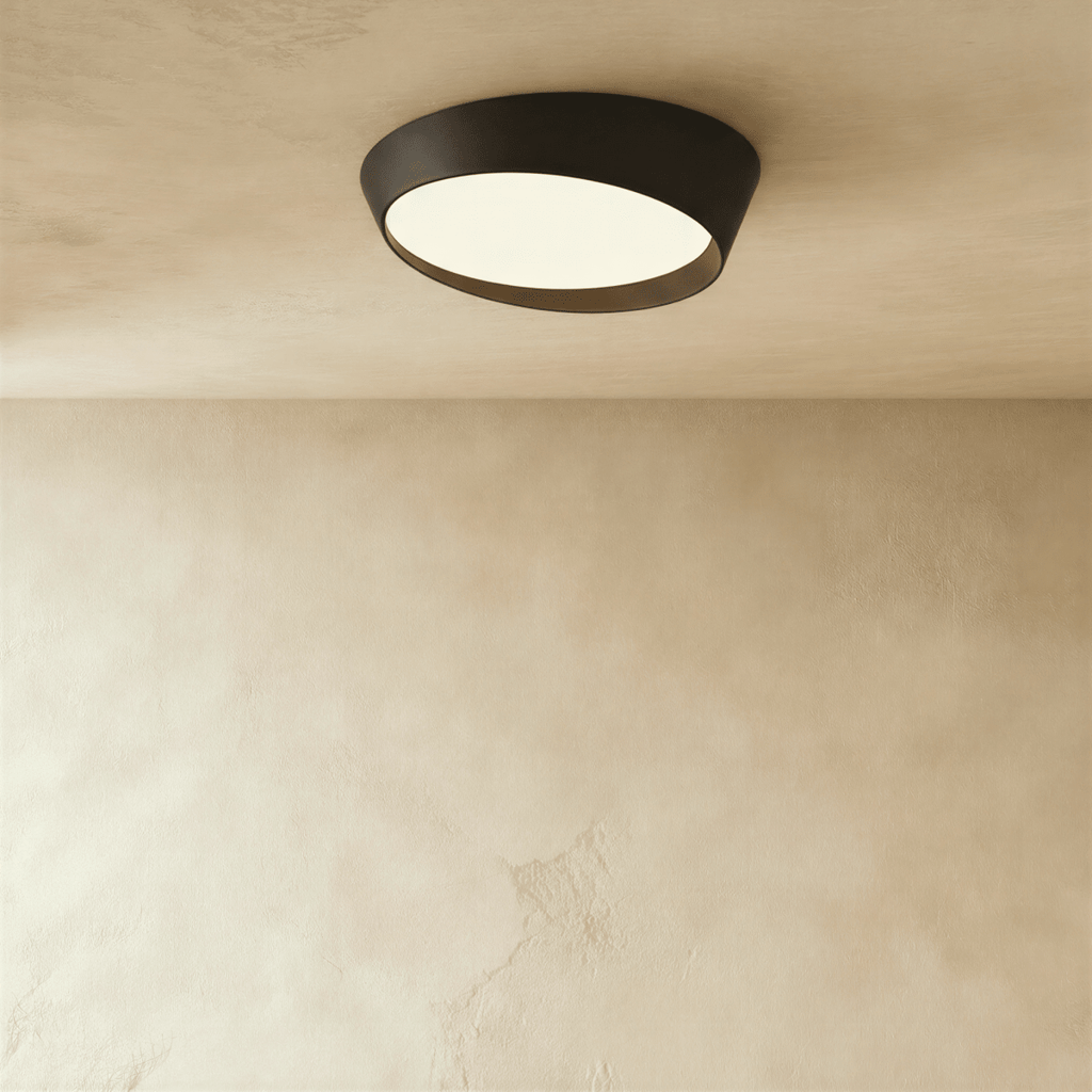 Clérachic black low-profile ceiling light with a slanted rim and a glowing frosted diffuser, seen from below on a limewash ceiling.