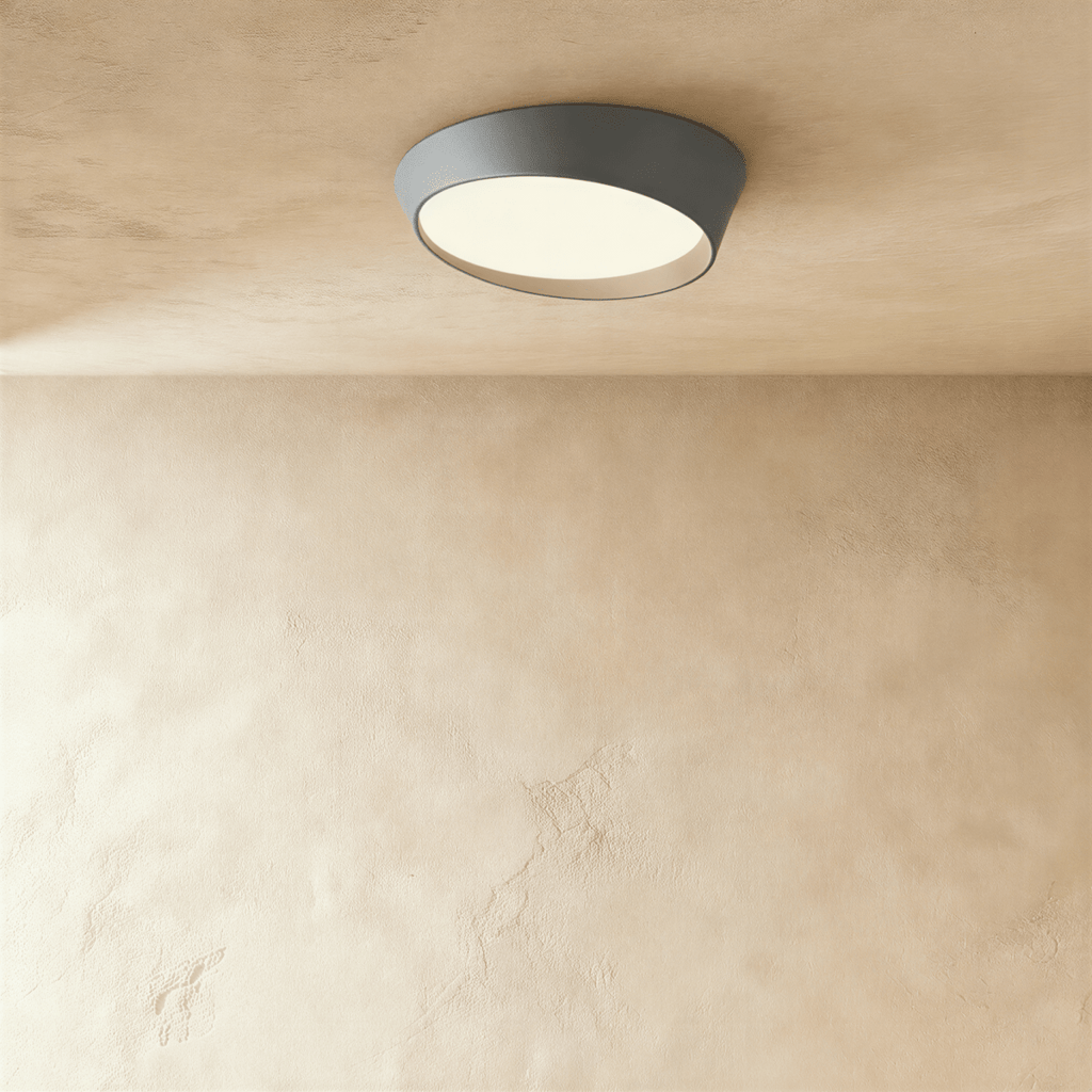 Clérachic round ceiling light in mid grey with a slanted rim and a warm-lit frosted diffuser, mounted on a limewash ceiling.