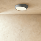 Clérachic round ceiling light in mid grey with a slanted rim and a warm-lit frosted diffuser, mounted on a limewash ceiling.