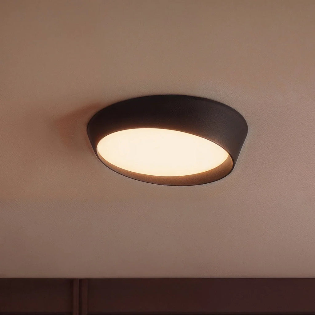 Clérachic black ceiling light close up, its slanted matte rim and warm frosted acrylic diffuser glowing against a pale ceiling.