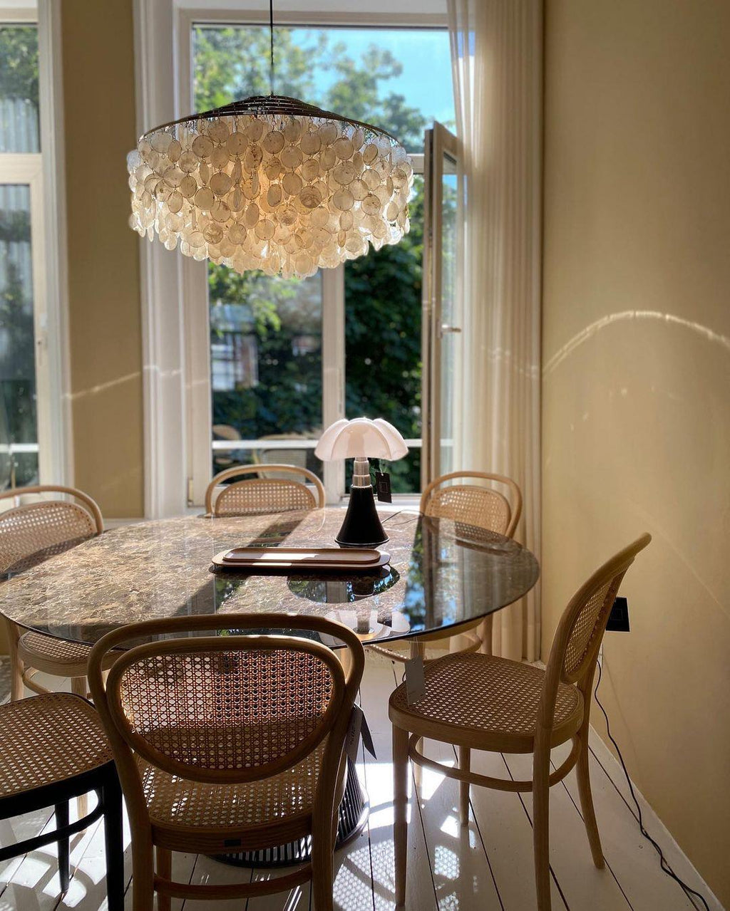 Clérachic shell chandelier above cane-backed chairs and a marble table beside a tall open window with garden views.
