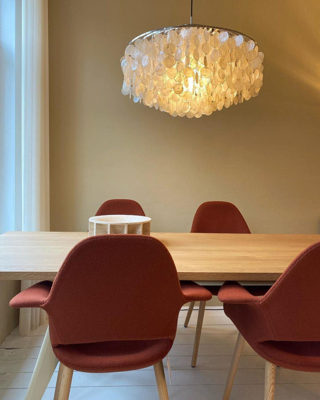 Lit Clérachic shell chandelier hanging above a pale oak table with rust-red upholstered chairs against a beige wall.