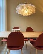 Lit Clérachic shell chandelier hanging above a pale oak table with rust-red upholstered chairs against a beige wall.