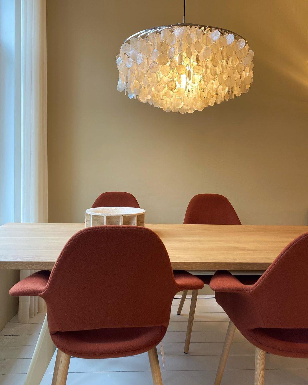 Lit Clérachic shell chandelier hanging above a pale oak table with rust-red upholstered chairs against a beige wall.