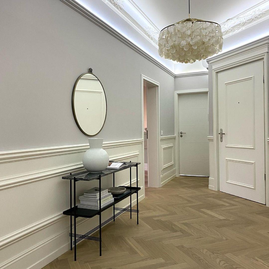 Clérachic shell chandelier lighting a hallway with a herringbone floor, white panelled walls and an oval mirror above a console.