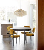 Gold-ringed Clérachic shell chandelier above a round black table and mustard velvet chairs, by a large window.
