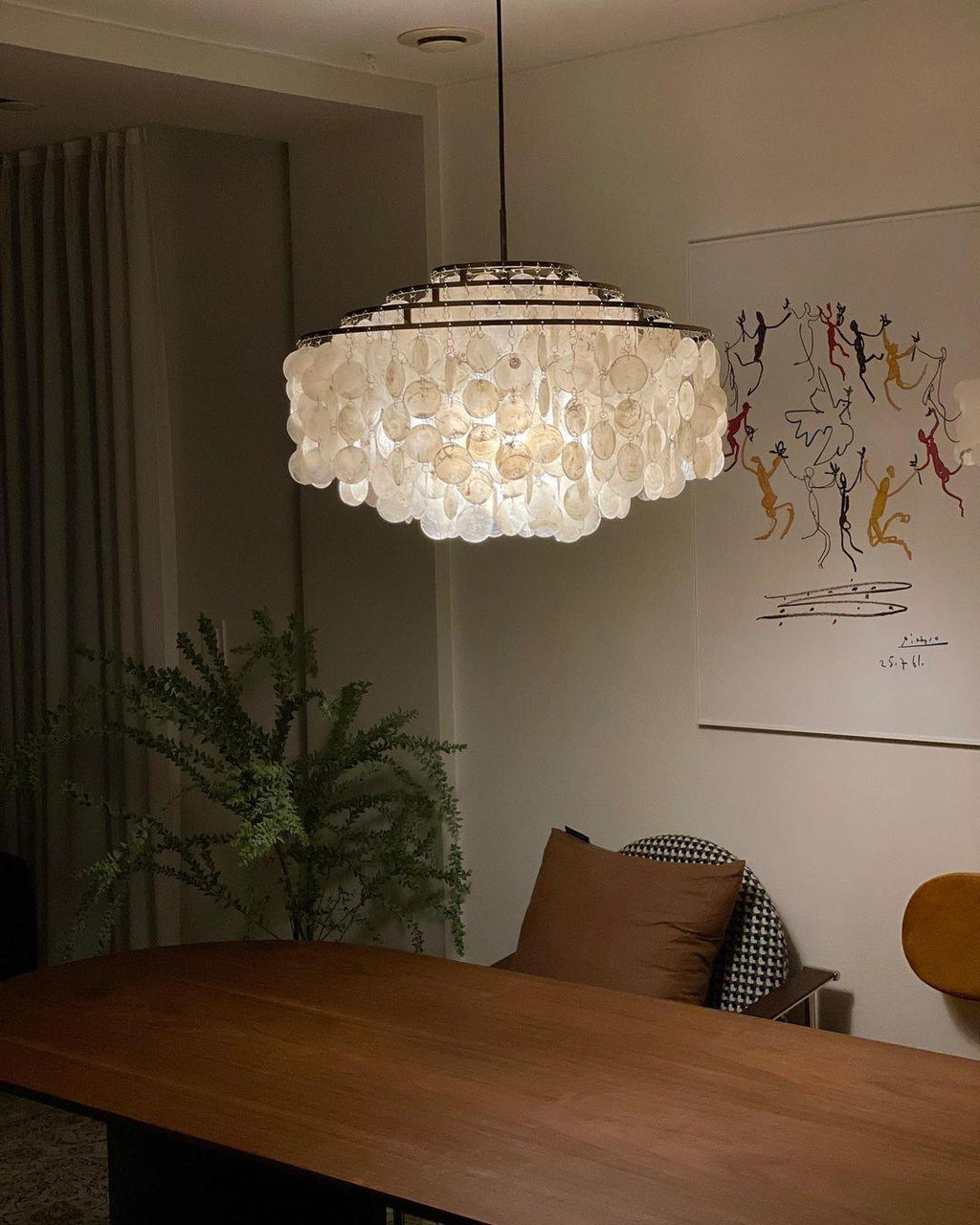 Closer view of a lit Clérachic shell chandelier over a walnut table, with a fern and a line-drawing print behind.