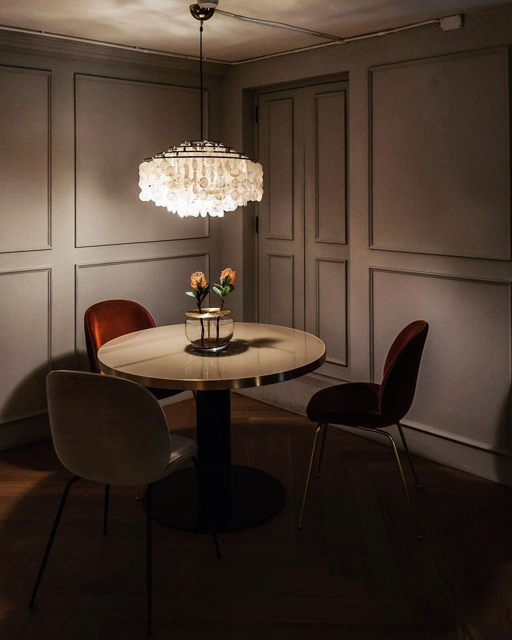 Lit Clérachic shell chandelier over a small round dining table in a dim grey-panelled room with velvet chairs.