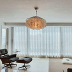 Clérachic shell chandelier glowing warm above a living room with a black leather lounge chair and ottoman by sheer curtains.