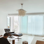 Clérachic shell chandelier, lit, hanging in a bright living room with a lounge chair and floor-length sheer curtains.