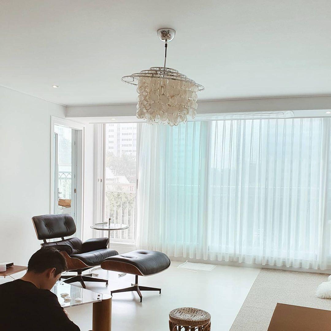 Clérachic shell chandelier, lit, hanging in a bright living room with a lounge chair and floor-length sheer curtains.