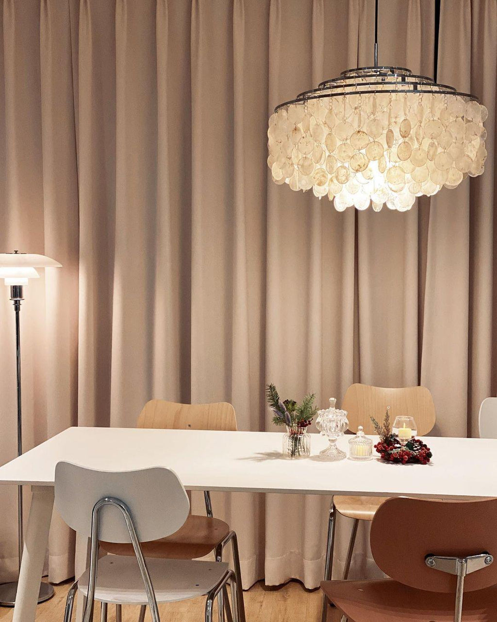 Clérachic shell chandelier with chrome rings hanging against taupe curtains, above a white table and bentwood chairs.