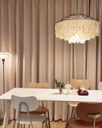 Clérachic shell chandelier with chrome rings hanging against taupe curtains, above a white table and bentwood chairs.
