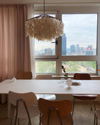 Clérachic shell chandelier in a tall window with a city skyline behind, above a long white table with wooden chairs.
