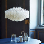 Large chrome-ringed Clérachic shell chandelier against a dark panelled wall, above a navy table set with glassware.