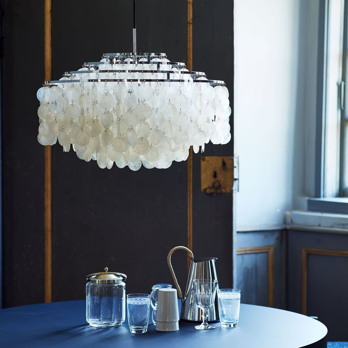 Large chrome-ringed Clérachic shell chandelier against a dark panelled wall, above a navy table set with glassware.