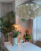 Part of a chrome-ringed Clérachic shell chandelier overhead, with a palm, a jug of wildflowers and a white table below.
