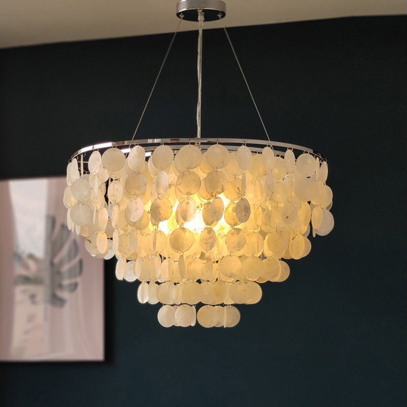 Lit Clérachic shell chandelier hung on fine cables against a deep teal wall, its discs glowing warm yellow.