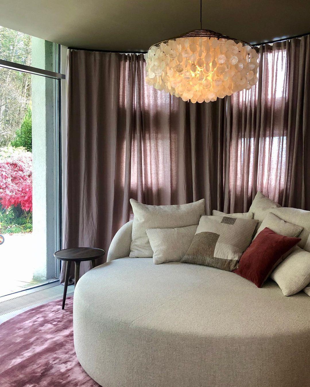 Clérachic shell chandelier glowing above a round cream sofa and mauve curtains in a curved window bay.