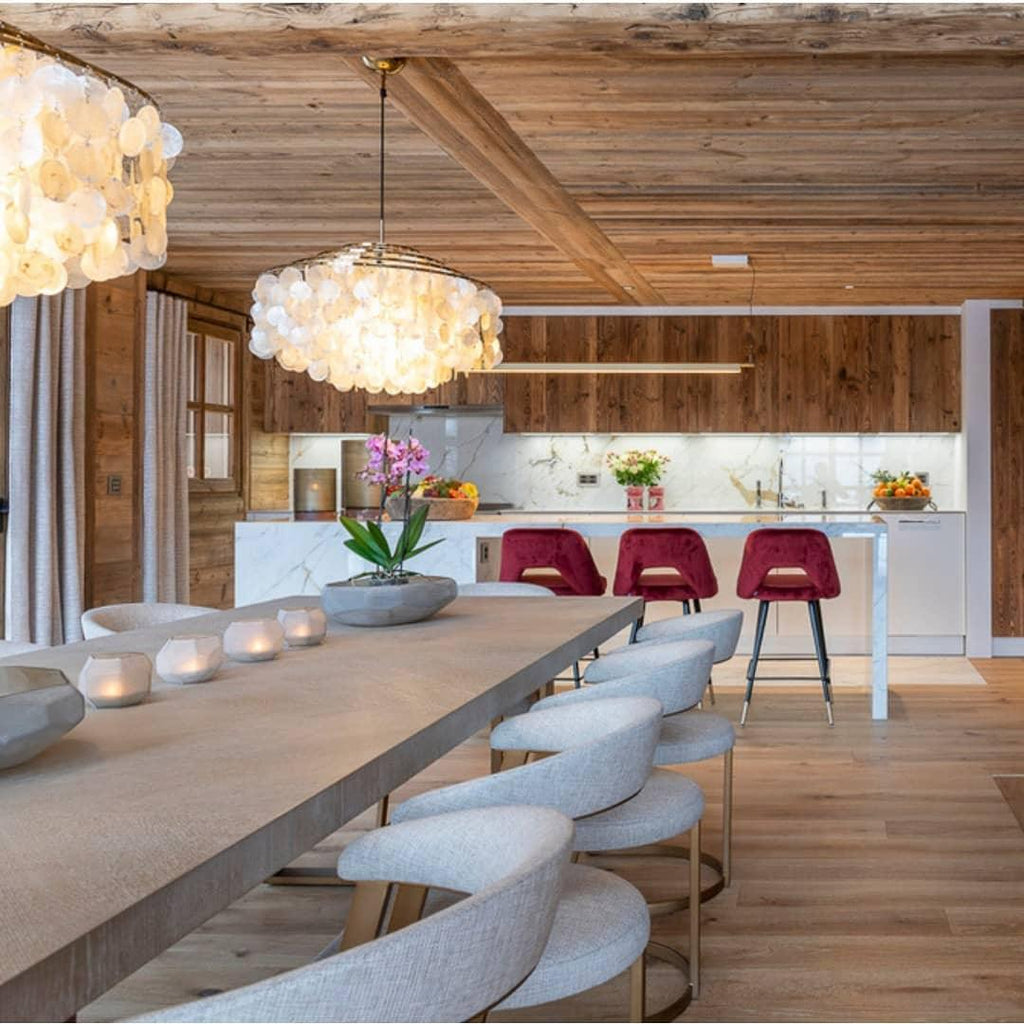 Clérachic shell chandelier over a long stone-topped table and boucle chairs, with a timber ceiling and kitchen beyond.
