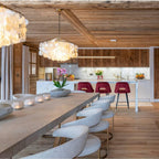 Clérachic shell chandelier over a long stone-topped table and boucle chairs, with a timber ceiling and kitchen beyond.