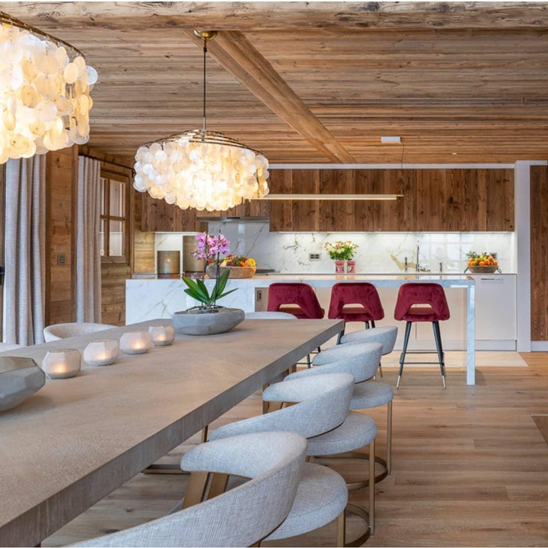 Clérachic shell chandelier over a long stone-topped table and boucle chairs, with a timber ceiling and kitchen beyond.