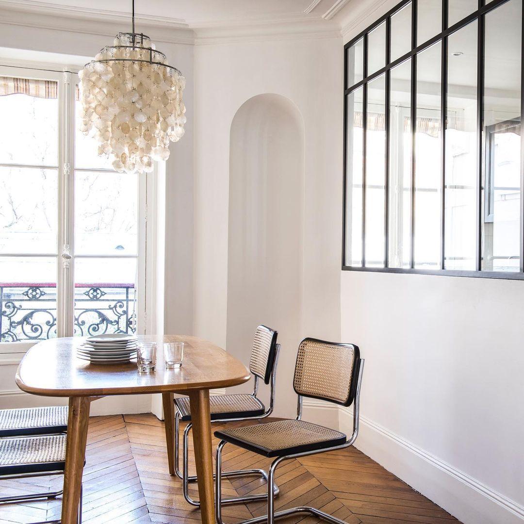 Clérachic shell chandelier in a Parisian dining room with a herringbone floor, a black-framed glass partition and cane chairs.