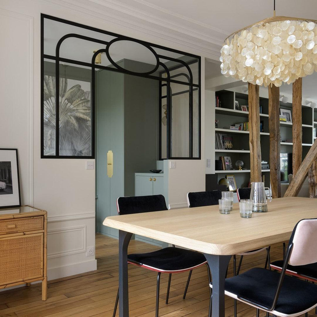Clérachic shell chandelier above a pale wood table and black velvet chairs, with timber posts and shelves behind.