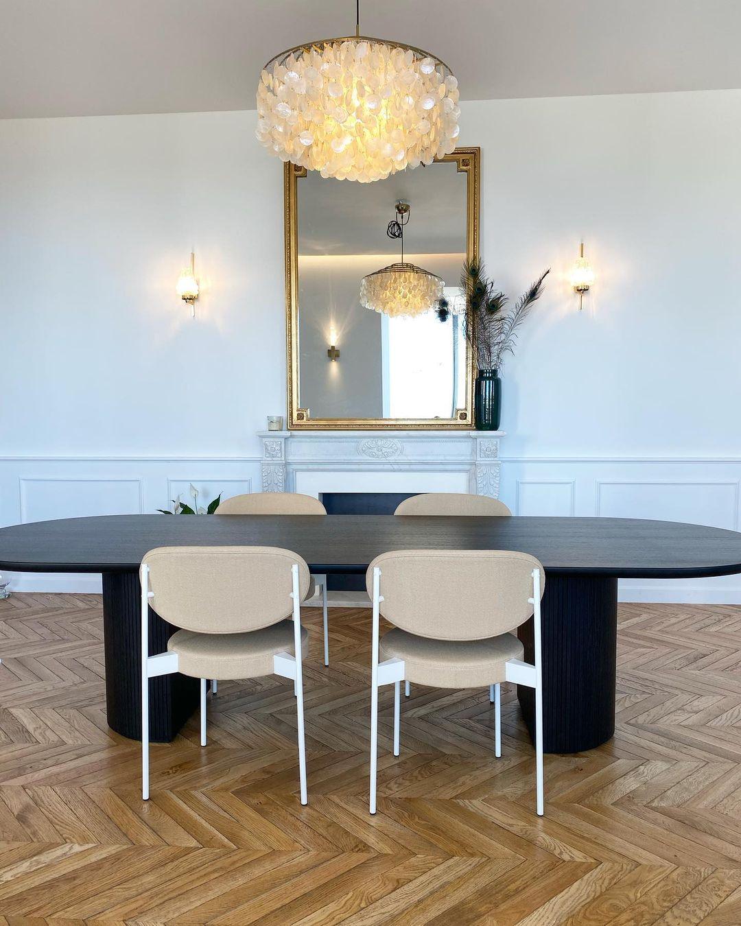 Clérachic shell chandelier over a long black oval table and cream chairs, with a gilt mirror and herringbone floor.