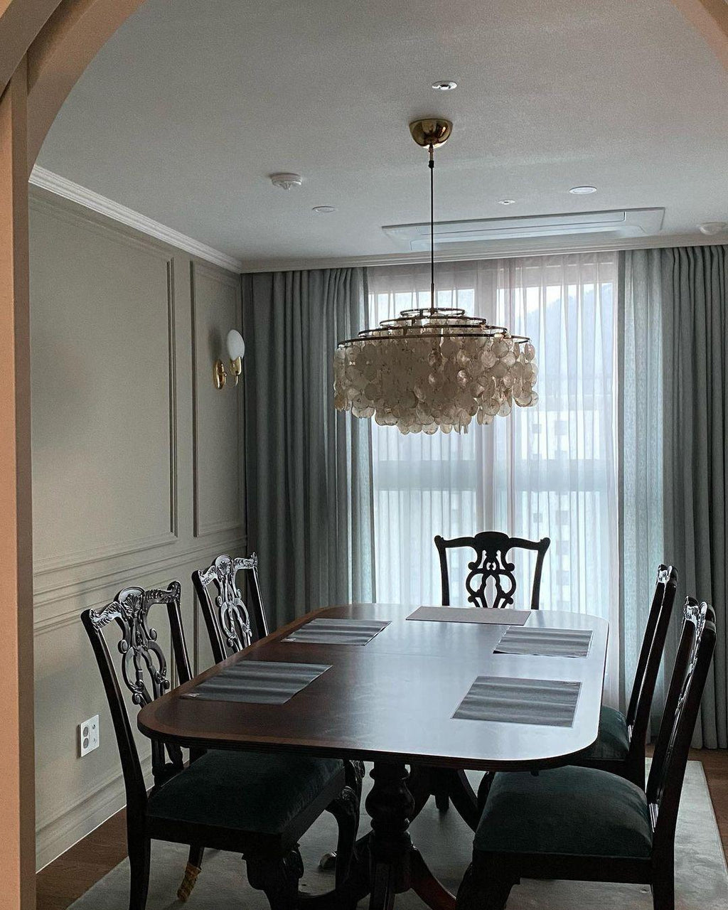 Clérachic shell chandelier above a dark wood dining table with carved chairs in a grey-green panelled room.