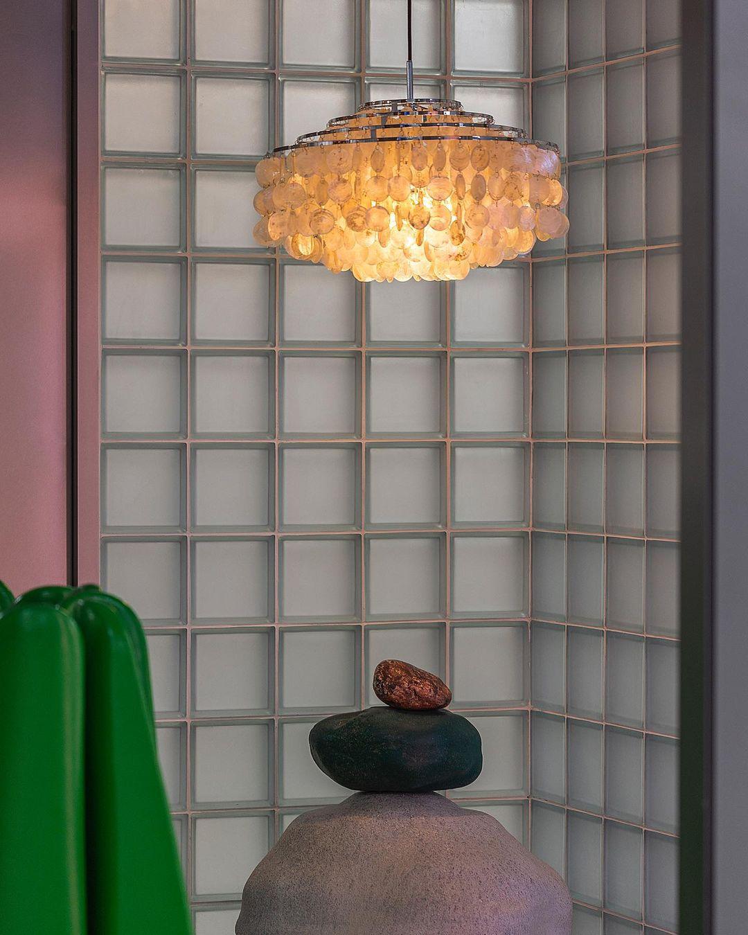 Lit Clérachic shell chandelier glowing amber in a glass-block alcove, with a green sculpture and stacked stones below.