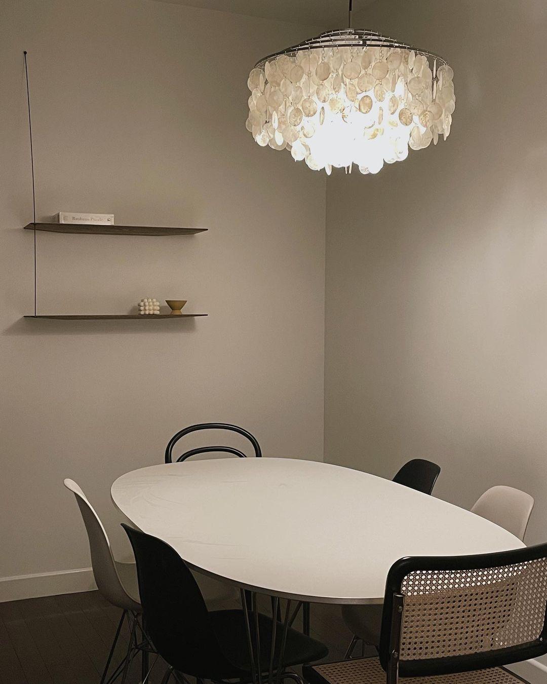 Clérachic shell chandelier above an oval white table with black chairs, in a corner with two slim wall shelves.
