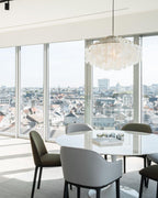 Clérachic shell chandelier by floor-to-ceiling windows overlooking rooftops, above a white round table and grey chairs.