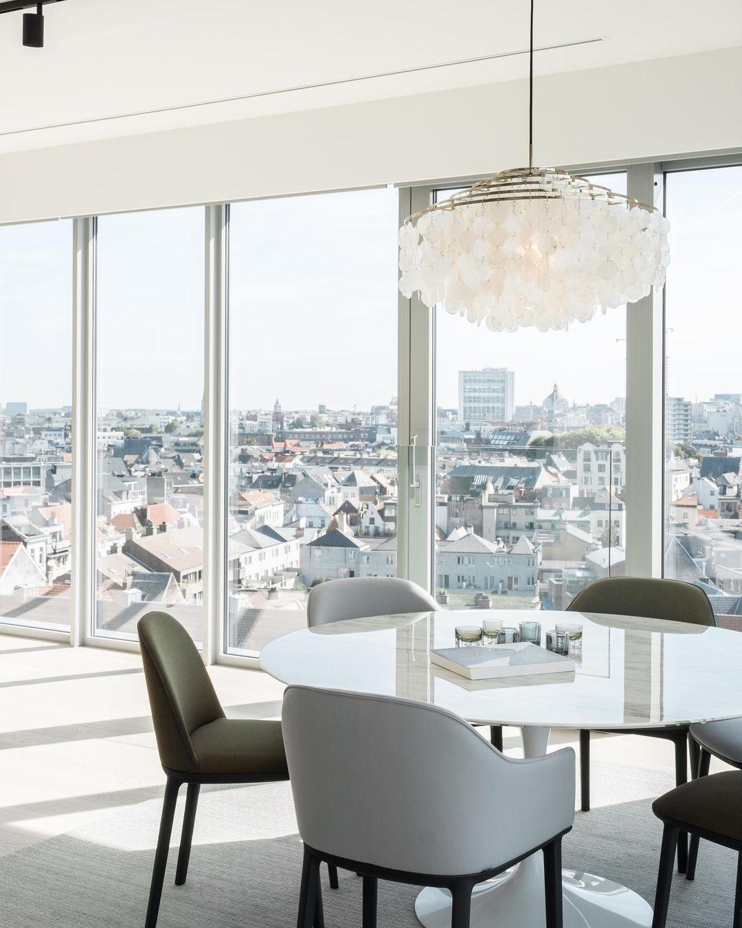 Clérachic shell chandelier by floor-to-ceiling windows overlooking rooftops, above a white round table and grey chairs.