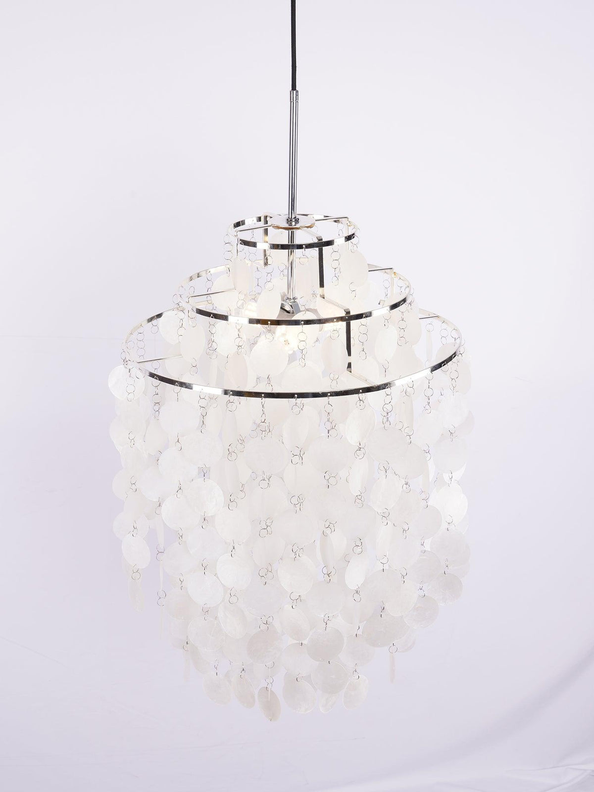 Narrow three-tier Clérachic shell chandelier with chrome rings, hung from a black cable against a pale lilac backdrop.