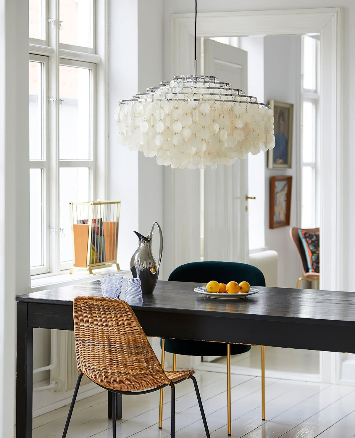 Clérachic shell chandelier above a black table with a wicker chair, by tall white windows and a pitcher on the table.