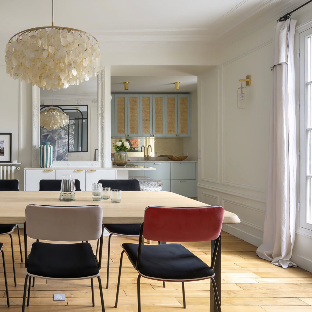 Clérachic shell chandelier at the top corner of a bright dining room with a pale wood table and red and grey chairs.