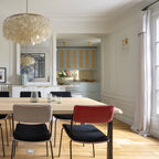 Clérachic shell chandelier at the top corner of a bright dining room with a pale wood table and red and grey chairs.